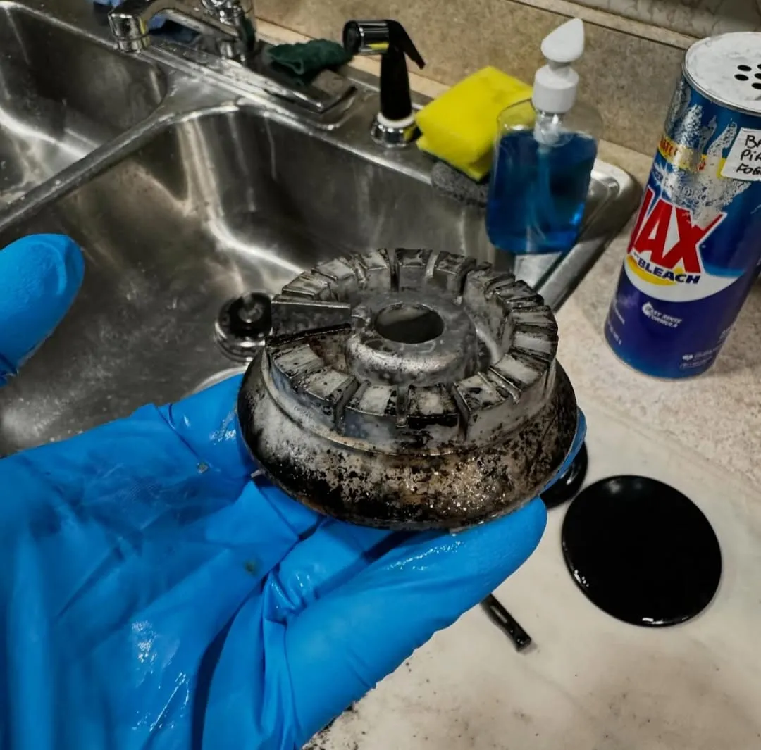 A gloved hand holding a greasy, charred stove burner cap over a sink, before scrubbing
