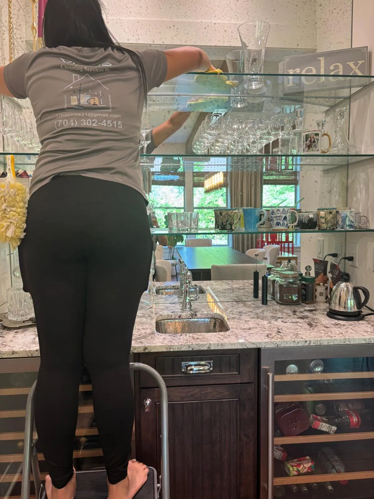 A cleaner on a step stool wiping glass shelves, seen from behind