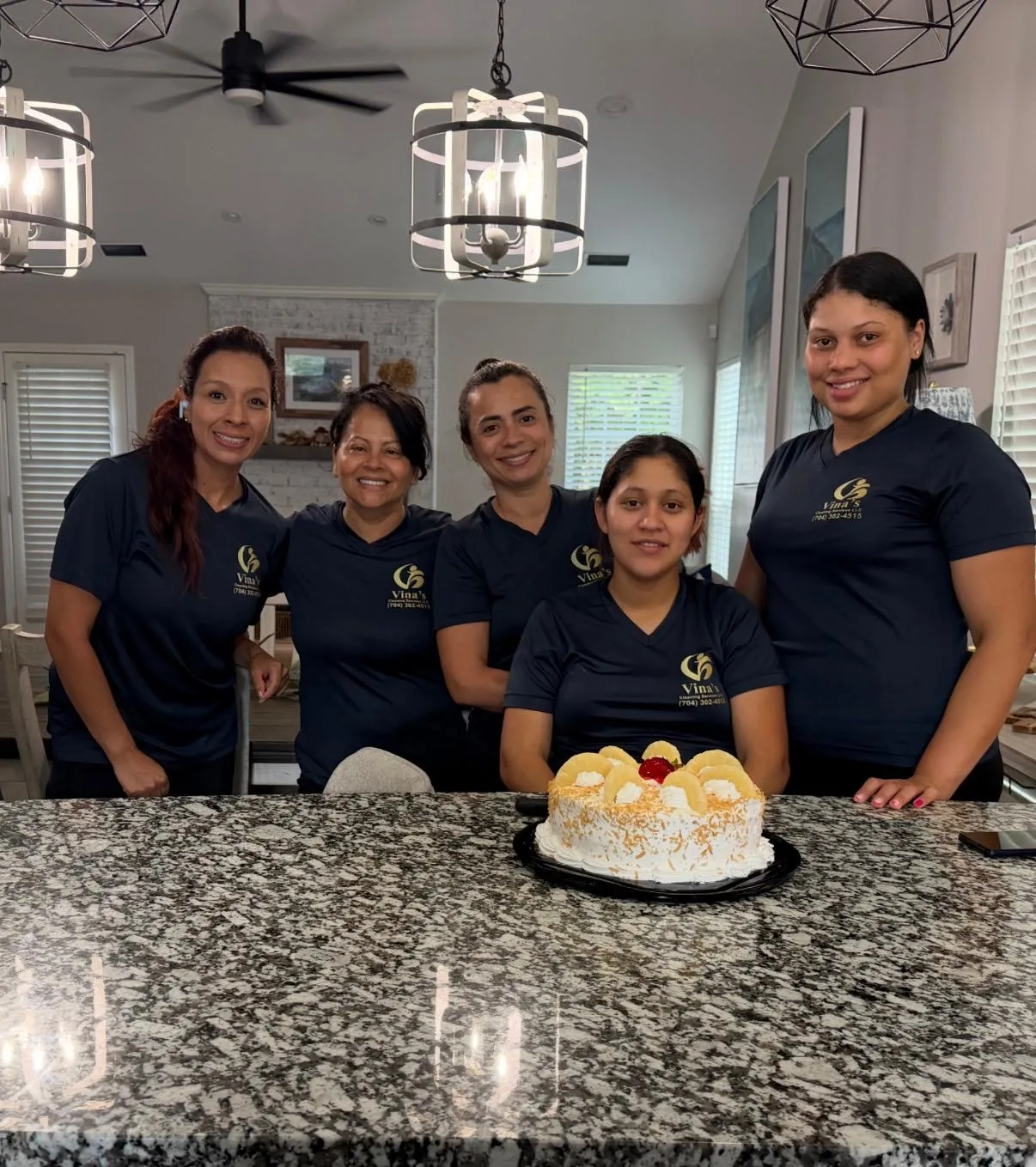Five members of the Vina's Cleaning Services team in navy polo shirts with the company logo, smiling around a kitchen island with a cake